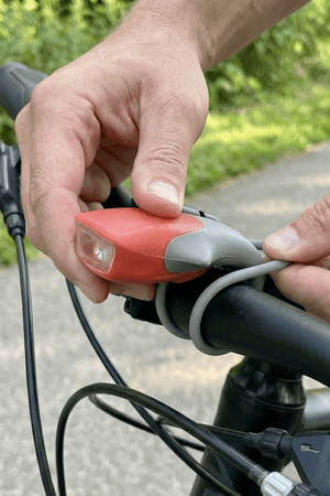 Hands fitting a red silicone LED bike light onto a handlebar with grey strap, outdoors - Optimum