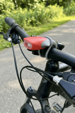 Red silicone LED bike light on bike handlebar along a leafy path, front view - Optimum
