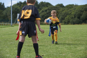 Young player running with ball in blue Optimum tag belt toward a defender in red flags, on grass
