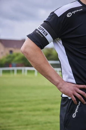 Black Optimum captain's armband worn on the arm of a player in black kit on a grass pitch - Optimum