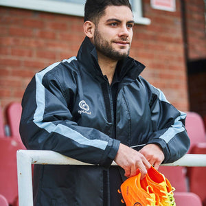 Man wearing black and grey Optimum sideline sub jacket holding orange boots, brick wall behind - Optimum