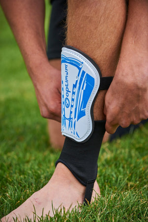 Blue and white Optimum Velocidade shin guard with black ankle sock and foot strap worn on leg on grass - Optimum