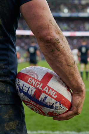 Muddy player holding a white and red Optimum England rugby ball with ENGLAND print in a stadium - Optimum