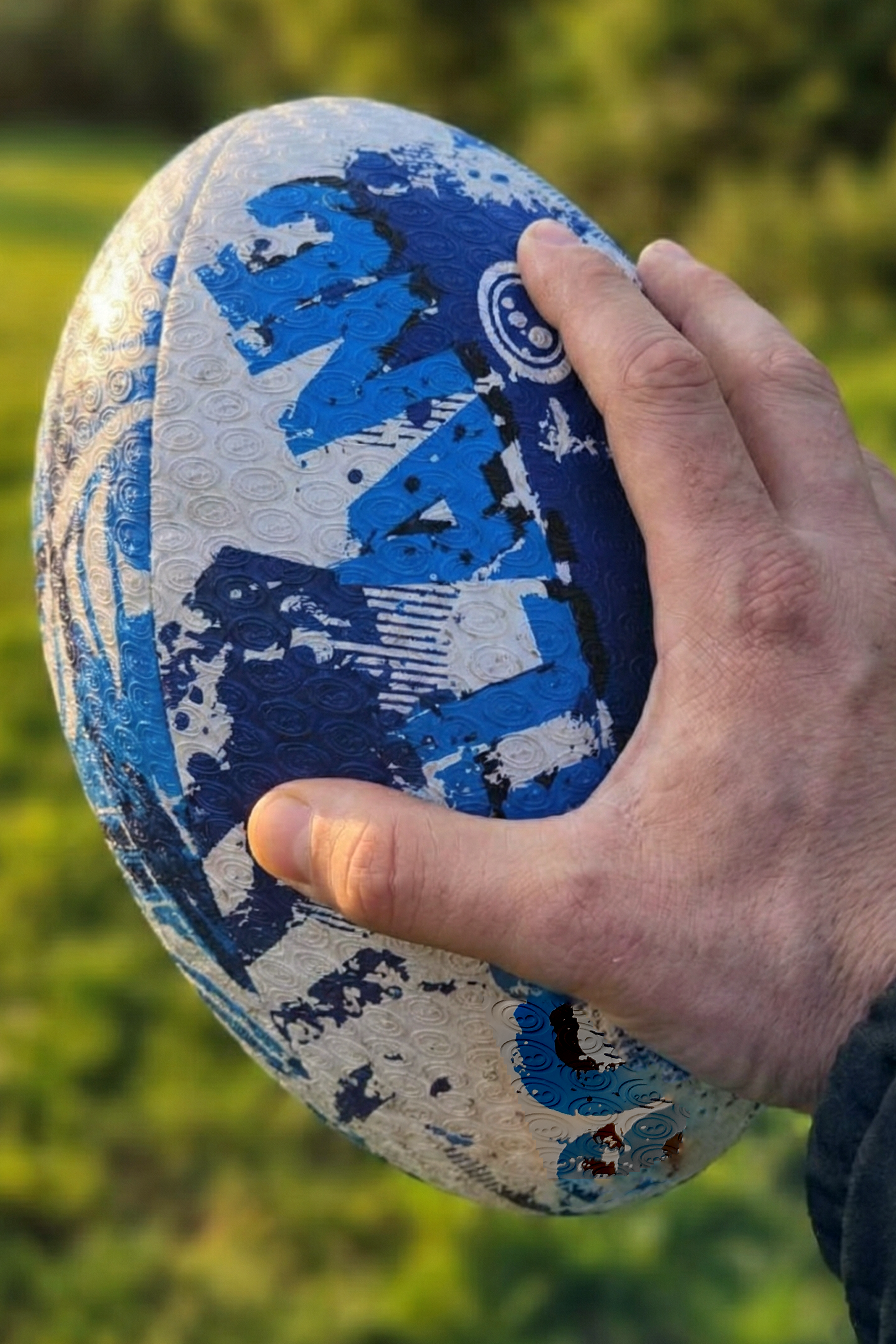 Hand holding a blue and white Optimum Scotland rugby ball outdoors with a grass background - Optimum