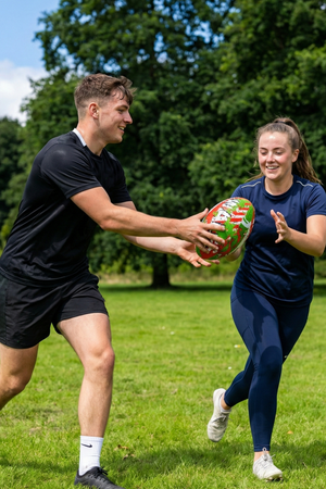 Two smiling players passing a red, green and white Optimum Wales rugby ball in a park - Optimum