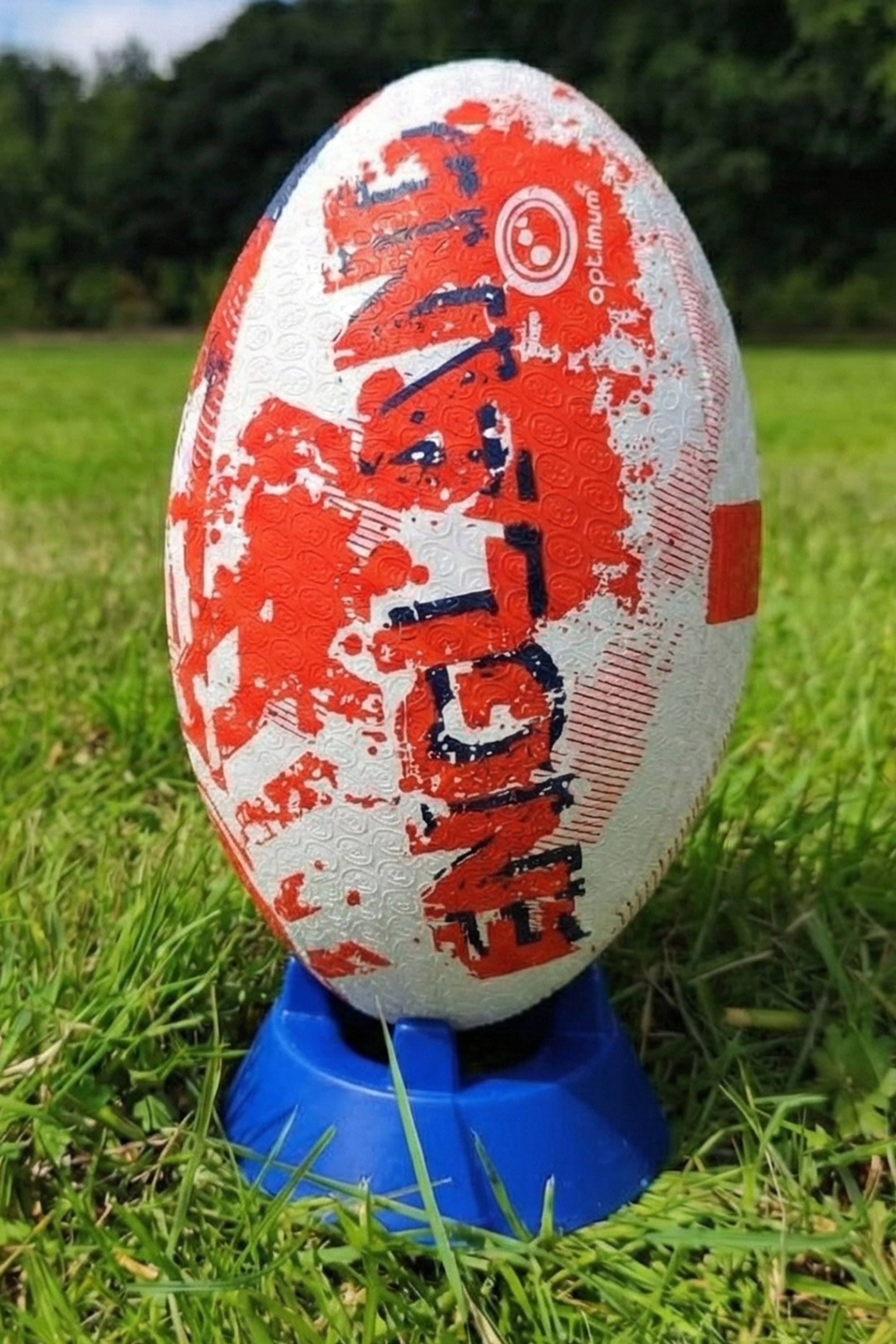 White and red Optimum England rugby ball with ENGLAND print upright on a blue kicking tee in grass - Optimum