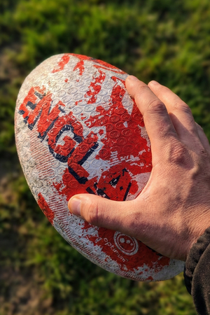 Hand holding a white and red Optimum England rugby ball with ENGLAND print above grass - Optimum