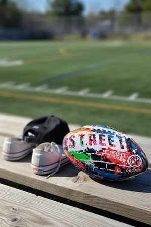 Multicolour Street American football with white laces on wooden bench, cap, trainers and sports field behind - Optimum