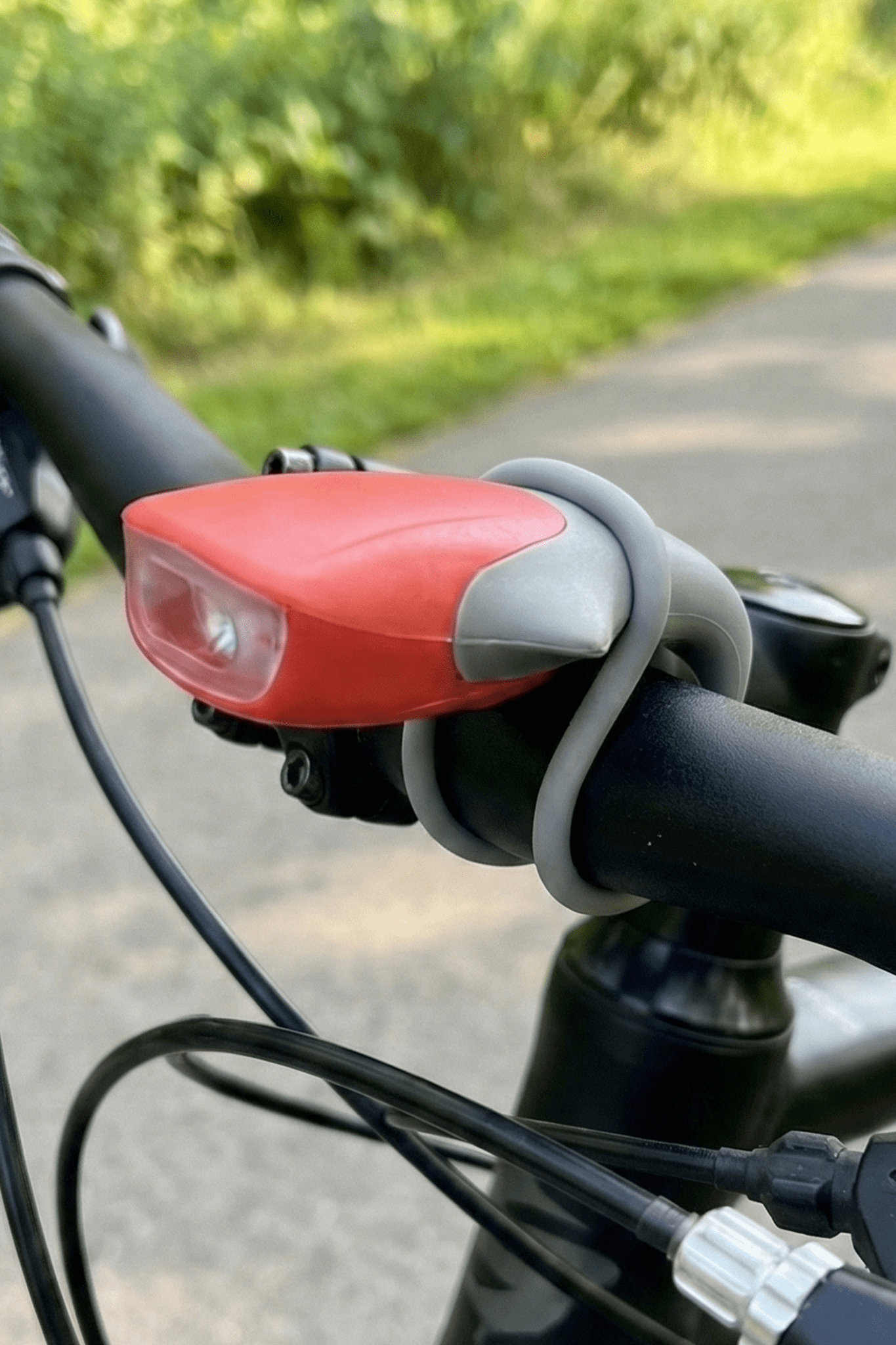 Red silicone LED bike light mounted on black handlebar with grey strap, outdoor path - Optimum
