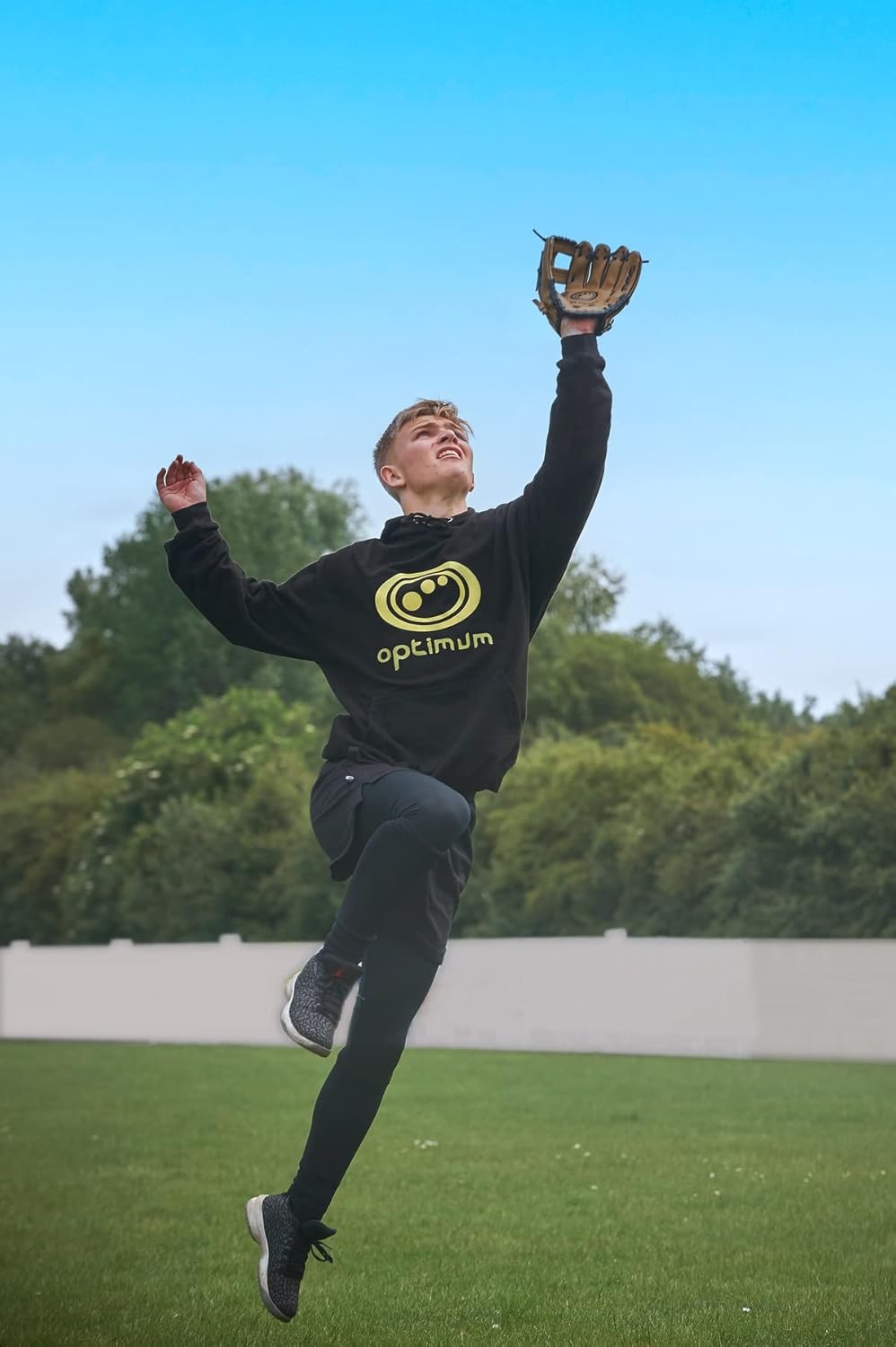 Player in black Optimum hoodie leaping to catch with tan baseball glove under blue sky - Optimum