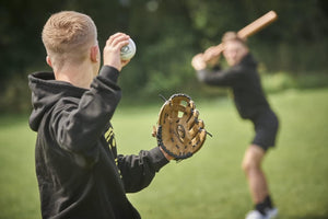 Pitcher in black hoodie with tan Optimum baseball glove and ball, batter behind on grass - Optimum