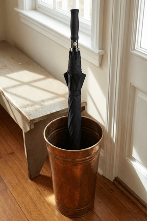 Closed black Optimum umbrella standing in copper stand by window indoors, lifestyle shot - Optimum