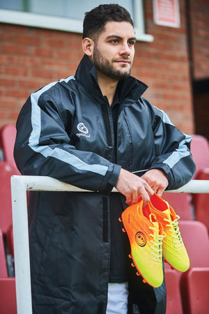 Man in black and grey Optimum sub jacket holding orange and yellow boots, brick wall behind - Optimum