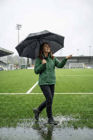 Woman holding open black Optimum umbrella on wet football pitch in rain, lifestyle shot - Optimum