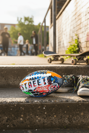 Multicolour graffiti-print Street American football with white laces on concrete steps, skateboard behind - Optimum