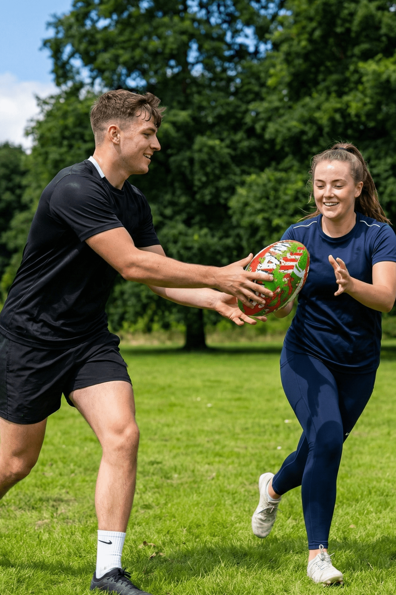 Two smiling players passing a red, green and white Optimum Wales rugby ball in a park - Optimum