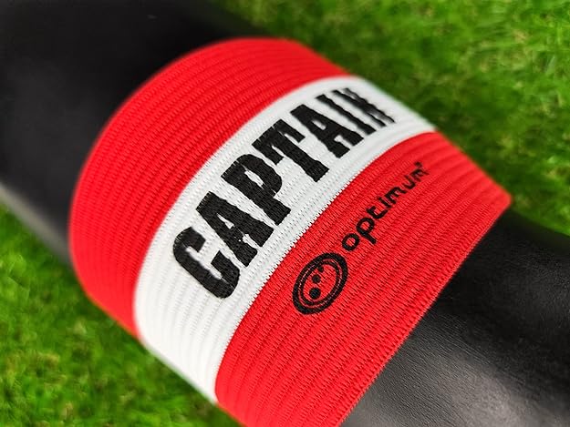 Red Optimum captain's armband with white CAPTAIN stripe and logo wrapped on a post on grass - Optimum