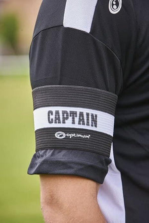 Close-up of black Optimum CAPTAIN armband worn on a player's arm over black kit on grass - Optimum