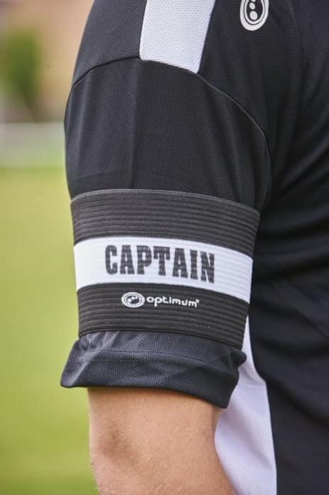 Close-up of black Optimum CAPTAIN armband worn on a player's arm over black kit on grass - Optimum