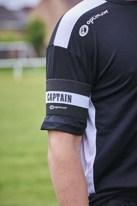 Black Optimum captain's armband worn over the sleeve of a player's black kit on a grass pitch - Optimum