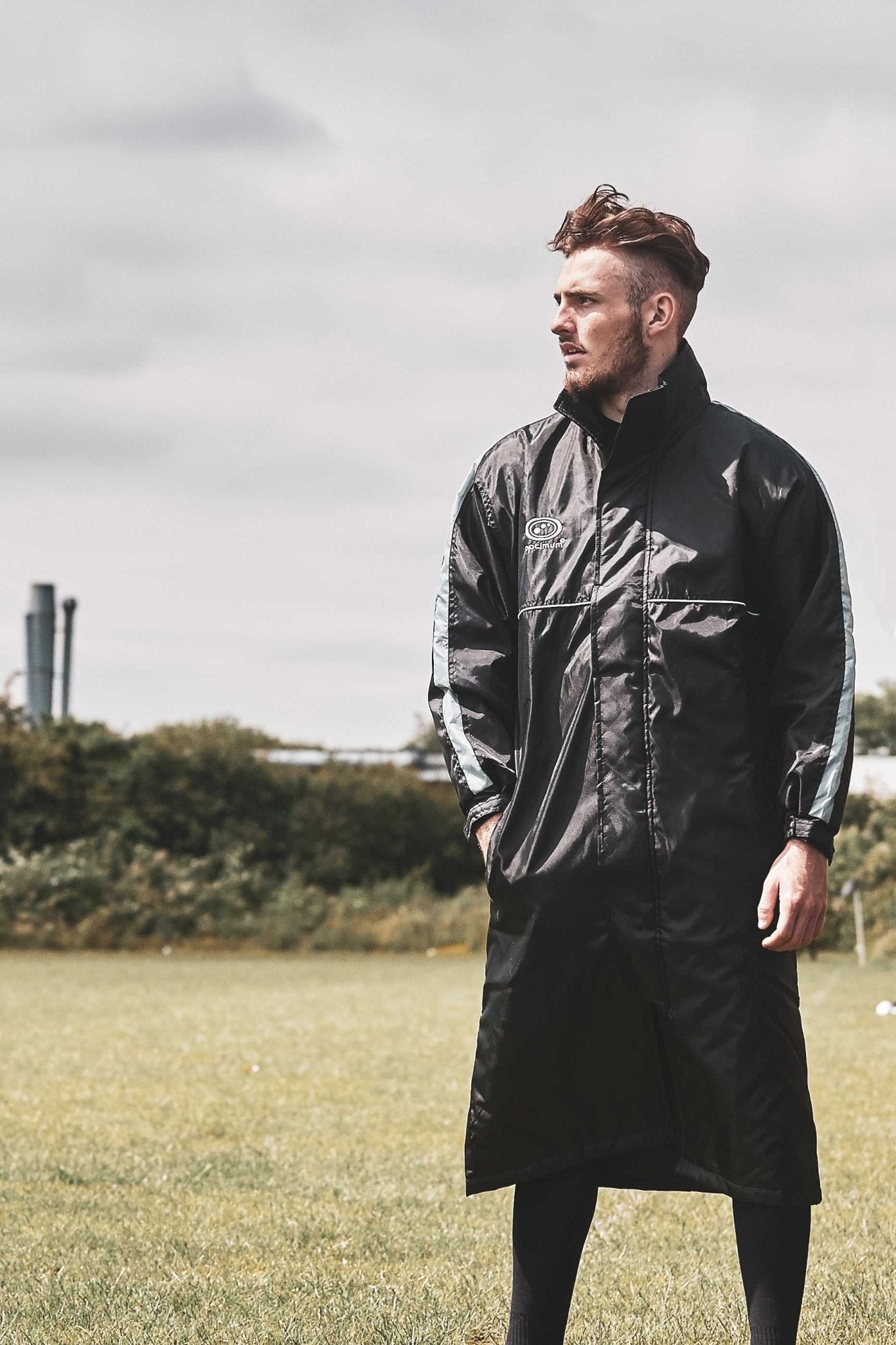 Man wearing long black Optimum sideline sub jacket standing on a grass field - Optimum