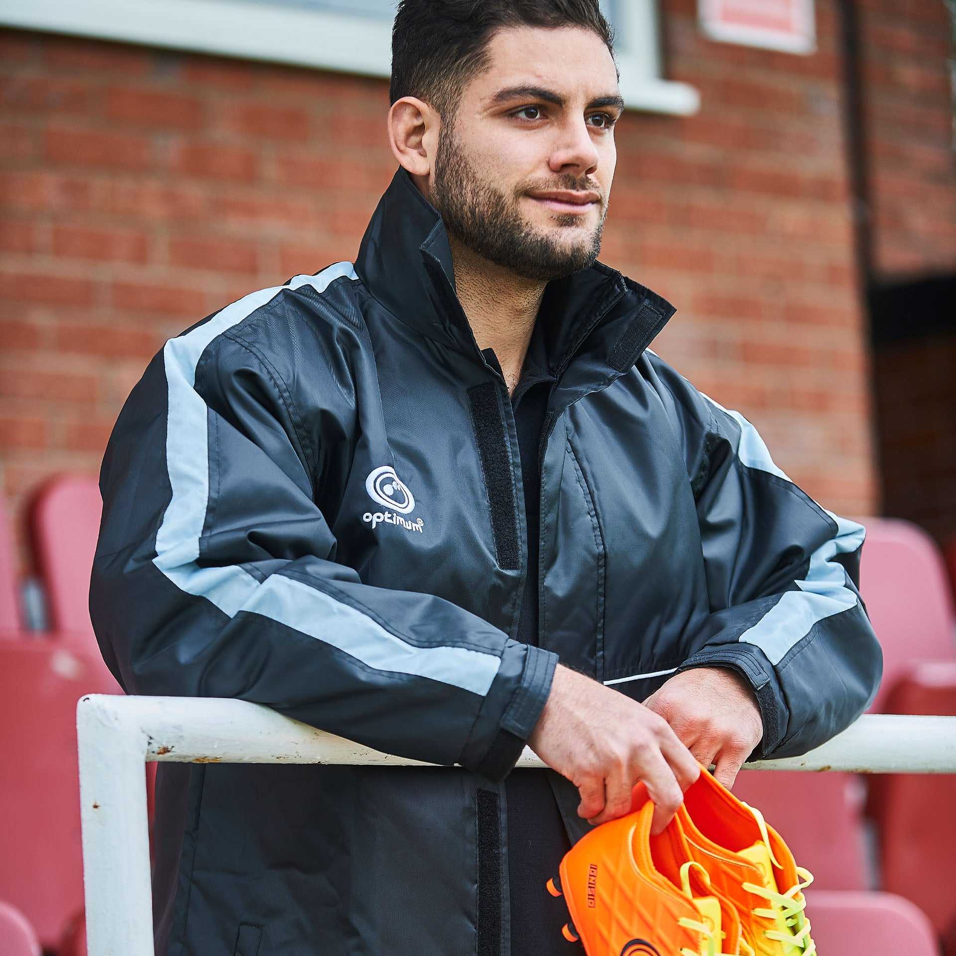 Man wearing black and grey Optimum sideline sub jacket holding orange boots, brick wall behind - Optimum