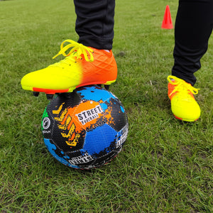 Foot in yellow and orange Optimum boot resting on the black street football on grass - Optimum