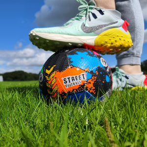 Multicolour Optimum street football on grass with a trainer resting on top, blue sky - Optimum