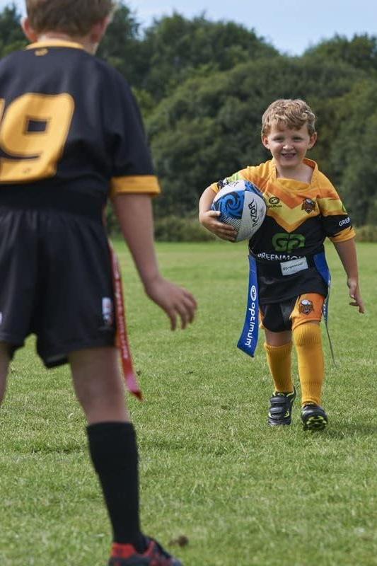 Young player running with ball in blue Optimum tag belt toward a defender in black kit, on grass