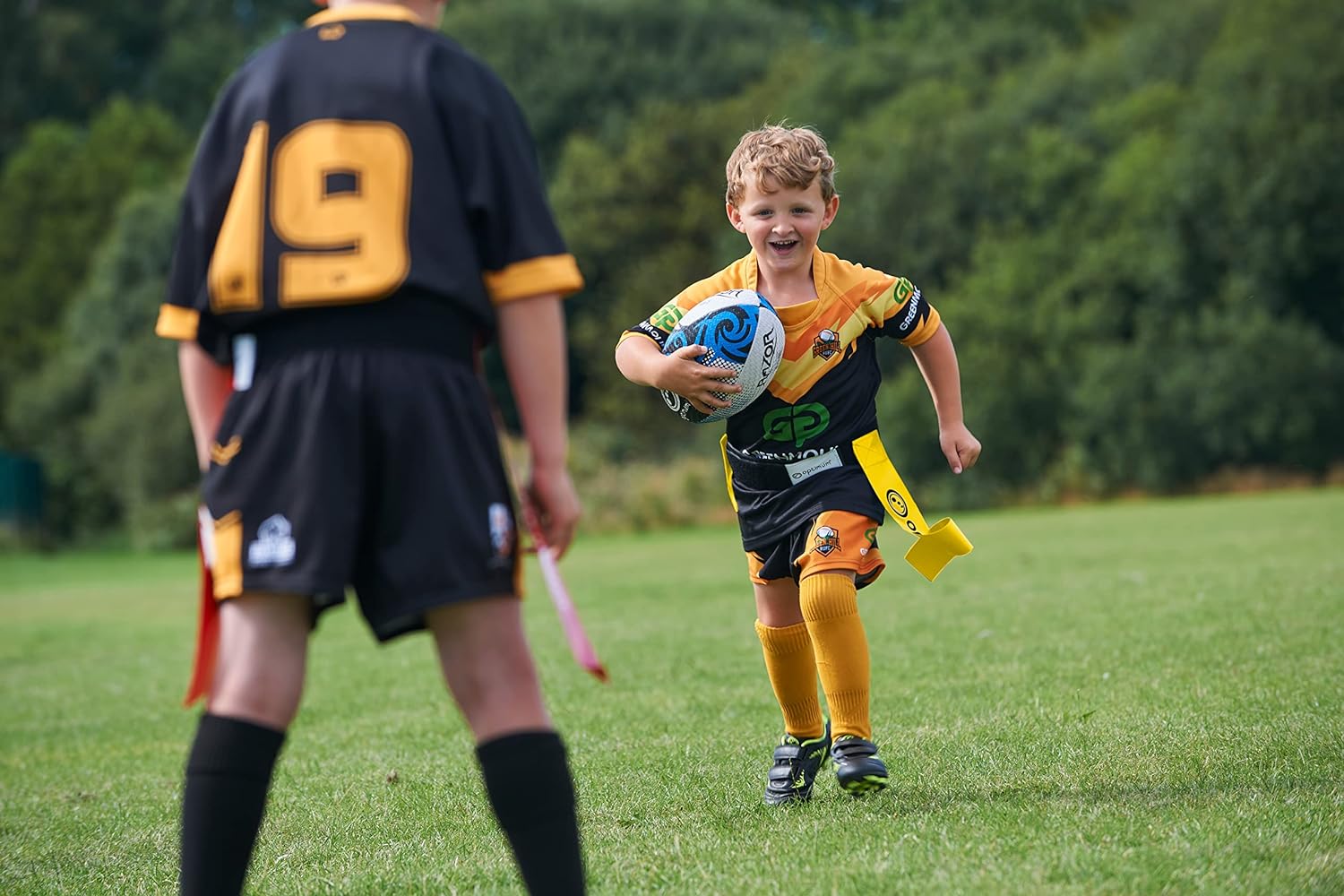 Young player running with ball in yellow Optimum tag belt toward a defender in red flags, on grass