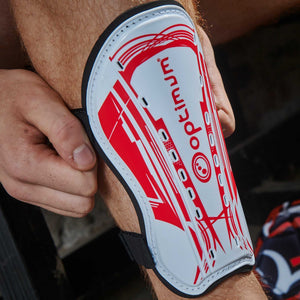 Hand holding white and red Optimum Velocidade shin guard with red print and logo against leg - Optimum