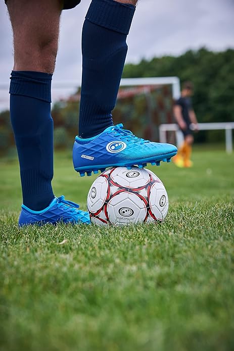 Player in blue Optimum Ignisio boots and navy socks resting foot on a football on a grass pitch - Optimum