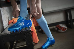 Player lacing up blue Optimum Ignisio football boots wearing light blue socks in a dark dressing room - Optimum