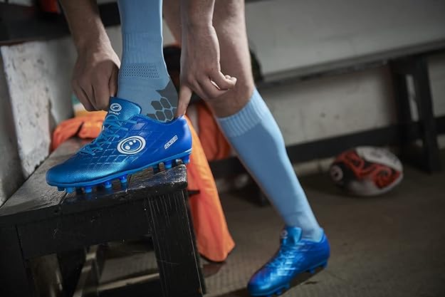 Player lacing up blue Optimum Ignisio football boots wearing light blue socks in a dark dressing room - Optimum