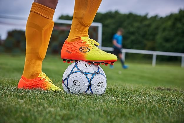 Player in orange Optimum Ignisio boots and orange socks resting foot on a football on a grass pitch - Optimum